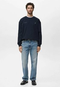 Man staat met zijn gezicht naar voren gekeerd in een marineblauwe sweatshirt, lichtblauwe jeans en zwarte schoenen tegen een effen witte achtergrond.