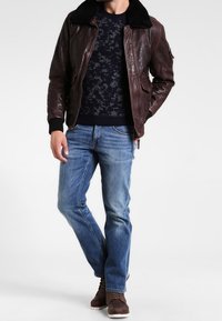 Braune Leder-Bomberjacke mit schwarzem Kunstpelzkragen, gemustertem dunklem Pullover, blauen Jeans und braunen Schnürstiefeln.