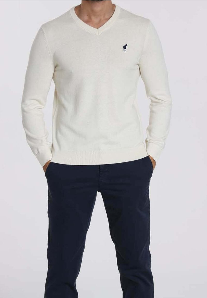 Homme portant un pull crème à manches longues avec col en V et petit logo de cheval noir sur la poitrine, ainsi qu'un pantalon bleu marine foncé, les mains dans les poches.