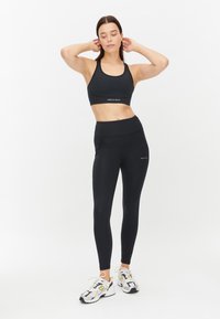 Zwarte sportbeha en high-waisted legging, beide gemaakt van rekbaar materiaal. Kenmerken subtiele logo-accenten en een naadloos ontwerp. Witte achtergrond.