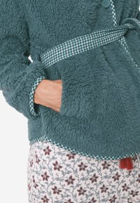 Türkiser, flauschiger Mantel mit grünem kariertem Krawatte und Besatz. Verfügt über eine weiche Textur und einen dekorativen roten Quasten am Saum. Weiße, blumenmusterte Hose ist sichtbar.