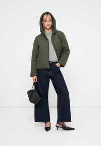 Giacca trapuntata verde oliva con cappuccio, t-shirt grigia, jeans scuri a gamba larga e décolleté nere, accompagnata da una borsa nera.