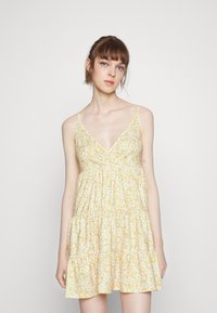 Vestido amarelo floral com decote em V, alças finas e saia em camadas. Apresenta um tecido leve com uma textura suave e pequenos padrões de flores brancas.
