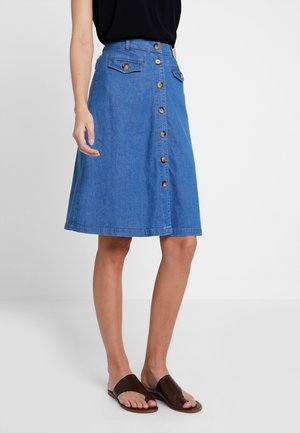 Femme portant une jupe en jean bleue à boutons sur le devant, longueur genou, un top noir sans manches et des sandales plates ouvertes marron foncé sur fond blanc.