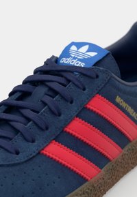 Marineblå ruskinds sneaker med røde striber, struktureret gummisål og blå tunge med Adidas-logoet og "MONTREAL" i guld.