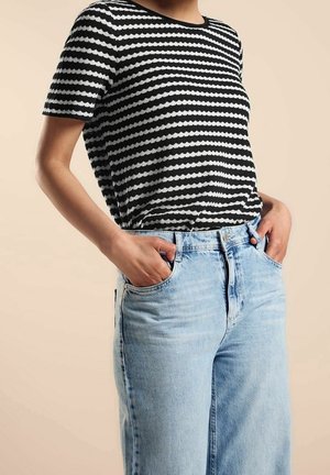 Personne portant un t-shirt rayé noir et blanc à manches courtes à bord festonné, rentré dans un jean taille haute bleu clair, les mains dans les poches.