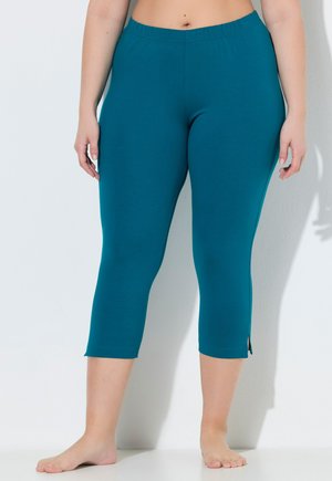 Person trägt türkisfarbene, gekürzte Leggings mit kleinen Seitenschlitzen und steht barfuß vor einer weißen Wand.