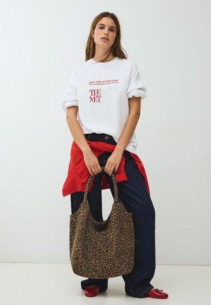 Fehér inget piros felirattal viselő nő, sötét farmerben, piros cipőben, derekán megkötött piros pulóverrel, kezében nagy leopárdmintás táskával.