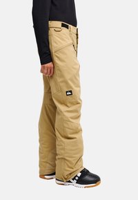 Personne portant un pantalon imperméable beige avec une poche latérale zippée et des bottes de neige noires avec semelles blanches et marron sur fond blanc.