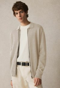 Hellbeige Reißverschluss-Cardigan mit subtiler Textur, getragen über einem weißen T-Shirt. Kombiniert mit hellen Hosen und einem schwarzen Gürtel mit silberner Hardware.