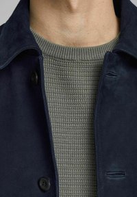 Námořnická suede bunda s knoflíkovým zapínáním, vrstvená přes strukturovaný, světle zelený pletený svetr s žebrovaným límcem.