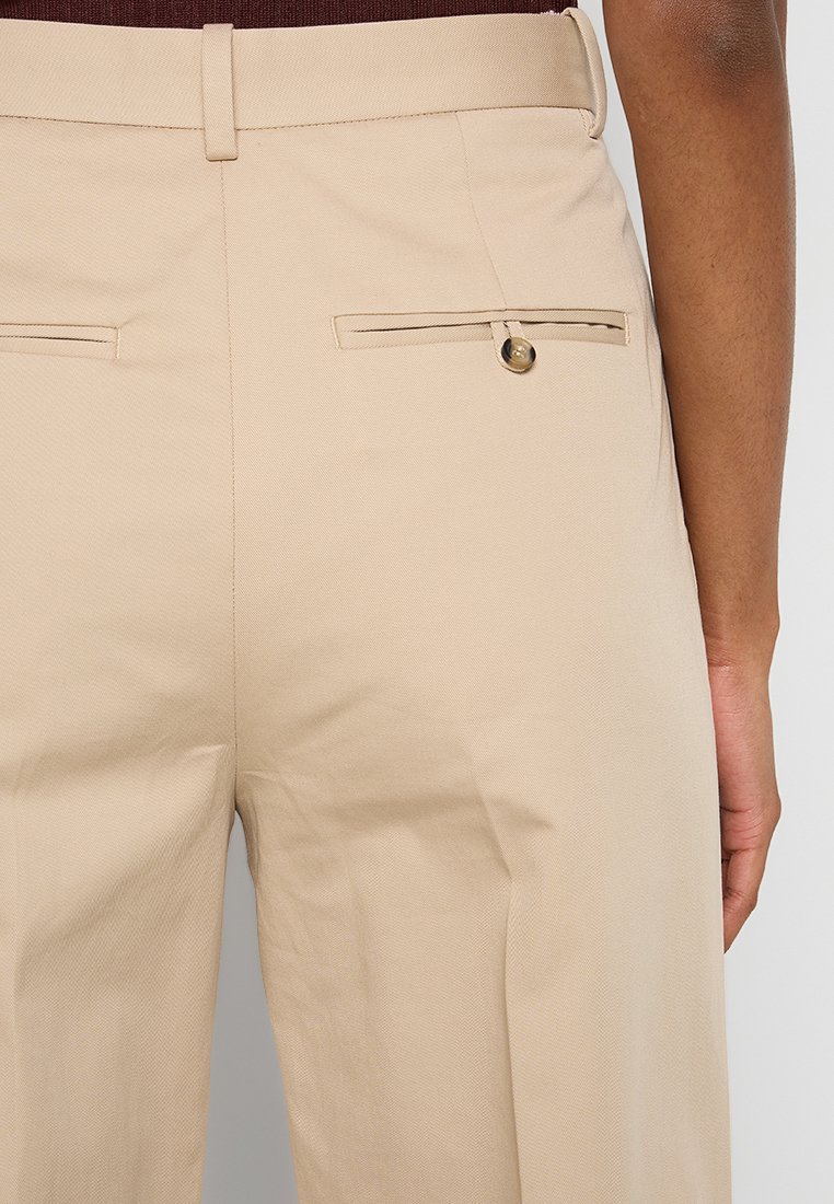 Pantaloni beige su misura con una texture liscia, due tasche posteriori con aperture orizzontali e dettagli in bottone color oro.