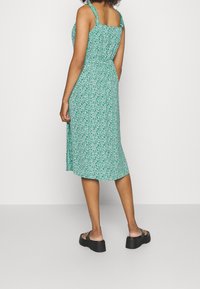 Vestido floral verde con tirantes ajustables, de longitud media y cintura elástica. Lleva sandalias de plataforma negras. Textura de tela suave.