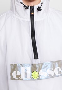 Vitt vindjacka med halvdragkedja fram, reflekterande patch med "ellesse"-logotyp och ett smiley-ansikte, detaljerad snörhuva.