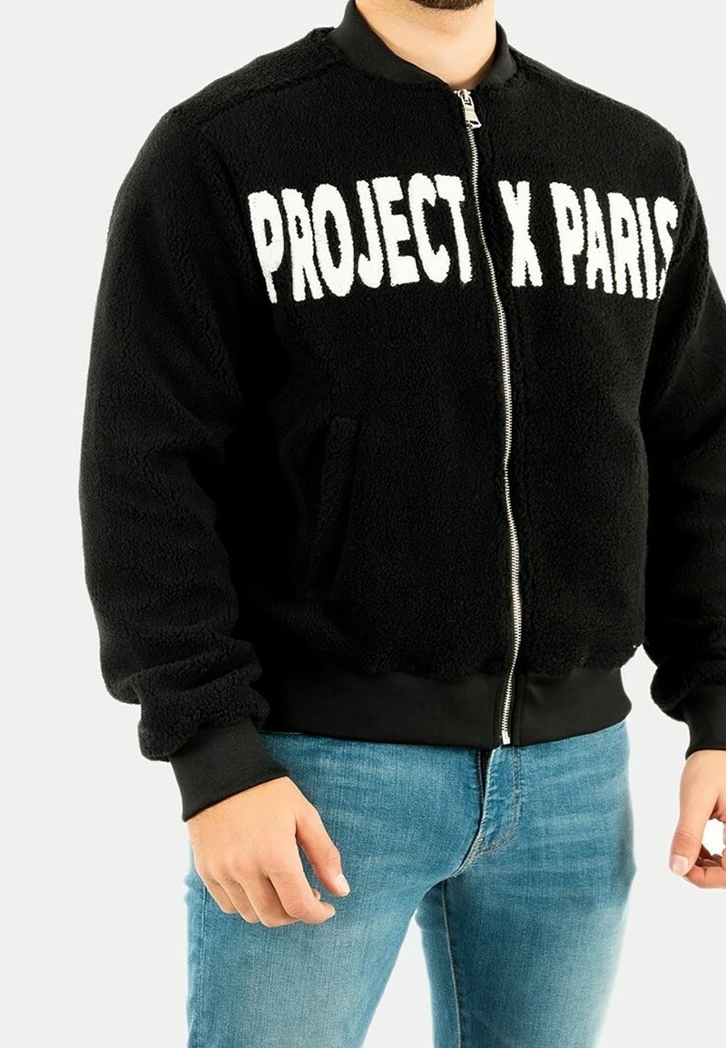 Man i svart zip-up fleecejacka med texten "PROJECT X PARIS" i vitt, tillsammans med blå jeans, mot en enfärgad bakgrund.