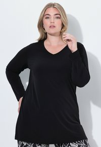 Schwarze langärmelige Tunika mit V-Ausschnitt, weichem Material und lockerer Passform. Das Modell trägt gemusterte Leggings mit blumigen Designelementen.