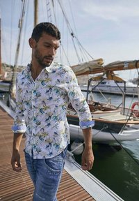Wit, met bloemenpatroon, overhemd met korte mouwen, met groene en blauwe bladeren, gecombineerd met blauwe jeans. Houten steiger en boten op de achtergrond.