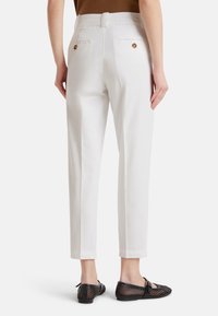 Donna che indossa pantaloni bianchi a vita alta con tasche posteriori con bottone e scarpe flat in rete nera, in piedi contro uno sfondo semplice.