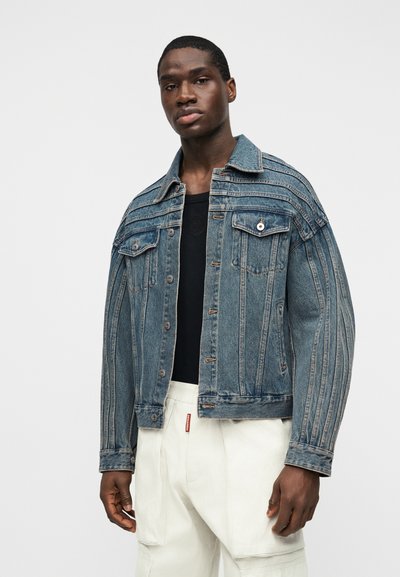 Άνδρας που φοράει μπλε τζιν μπουφάν με κάθετες ραφές, μαύρο πουκάμισο και λευκό παντελόνι, στέκεται μπροστά σε μια απλή λευκή επιφάνεια.