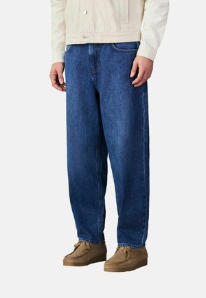 Mand iført løstsiddende blå denimjeans, beige mokkasin-sko i ruskind og en cremefarvet jakke, stående foran en ensfarvet baggrund.