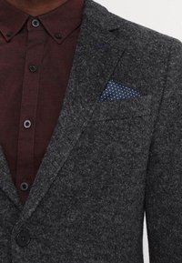 Blazer grigio scuro con texture sopra una camicia bordeaux abbottonata, con fazzoletto da taschino blu navy decorato con piccole stelle bianche nella tasca del petto del blazer.