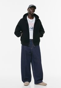 Zwarte hoodie met een rits aan de voorkant, grijze pet, witte T-shirt met grafisch ontwerp en losse blauwe spijkerbroek. Lichte grijze sneakers maken de look compleet.