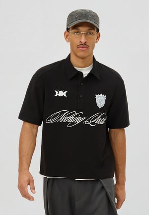 Férfi fekete polosben, fehér "Nothing Lasts" felirattal, fekete sapkában, szemüvegben és szürke nadrágban, egyszínű háttér előtt.