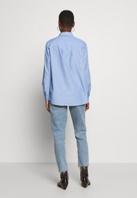 Lichtblauw overhemd met een ontspannen pasvorm, lange mouwen en een rugplooi. G gedragen met lichtblauwe jeans en donkere enkellaarsjes.