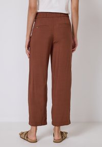Bruine, wijde pantalons met een elastische tailleband. Voorzien van twee achterzakken en een gladde textuur. Gecombineerd met platte, beige sandalen.