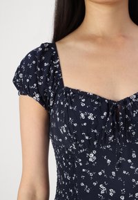 Námořnické modré šaty s krátkými nafouklými rukávy, květinovým vzorem, čtvercovým výstřihem, smockovaným živůtkem a vázáním na přední straně. Měkká textura látky.