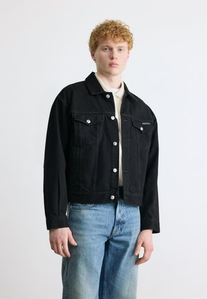 Fiatal férfi göndör vörös hajjal fekete farmerdzsekit visel világos ing felett és kék farmert, egy sima fehér háttér előtt áll.