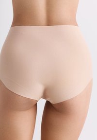 Vrouw in naadloze huidkleurige hoge taille onderbroek, gezien vanaf de achterkant tegen een effen lichte achtergrond.