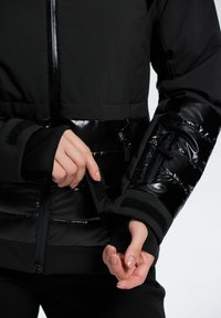 Sort pufferjakke med blanke paneler, lynlåslommer og velcro-lukninger ved håndleddene. Har en glat mat finish og struktureret design.