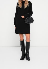 Abito nero a maniche lunghe abbinato a stivali in pelle nera al ginocchio e a una piccola borsa rigida con motivo trapuntato.