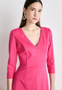 Fuchsia dress with three-quarter sleeves, V-neckline, structured bodice, and seam details. Model wears large gold hoop earrings.