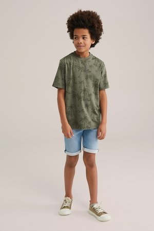 Oliven grøn kortærmet skjorte med palme-mønster, kombineret med lyse denimshorts og grønne sneakers. Afslappet sommeroutfit.