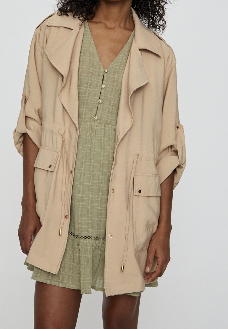 Beige oversized jacket with rolled sleeves, multiple front pockets, and gold accents, layered over a light green, buttoned, checkered dress.