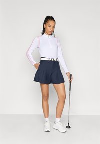 Valkoinen pitkähiha toppi, jossa on vetoketju ja vaaleanpunaisia yksityiskohtia, yhdistettynä tummansiniseen laskostettuun golfhameeseen ja valkoisiin urheilukenkään, pidellen golfmailaa.