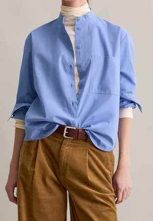 Persoon draagt een lichtblauw overhemd met knopen over een beige coltrui, met bruine corduroy broek en een bruine riem, staand tegen een effen achtergrond.
