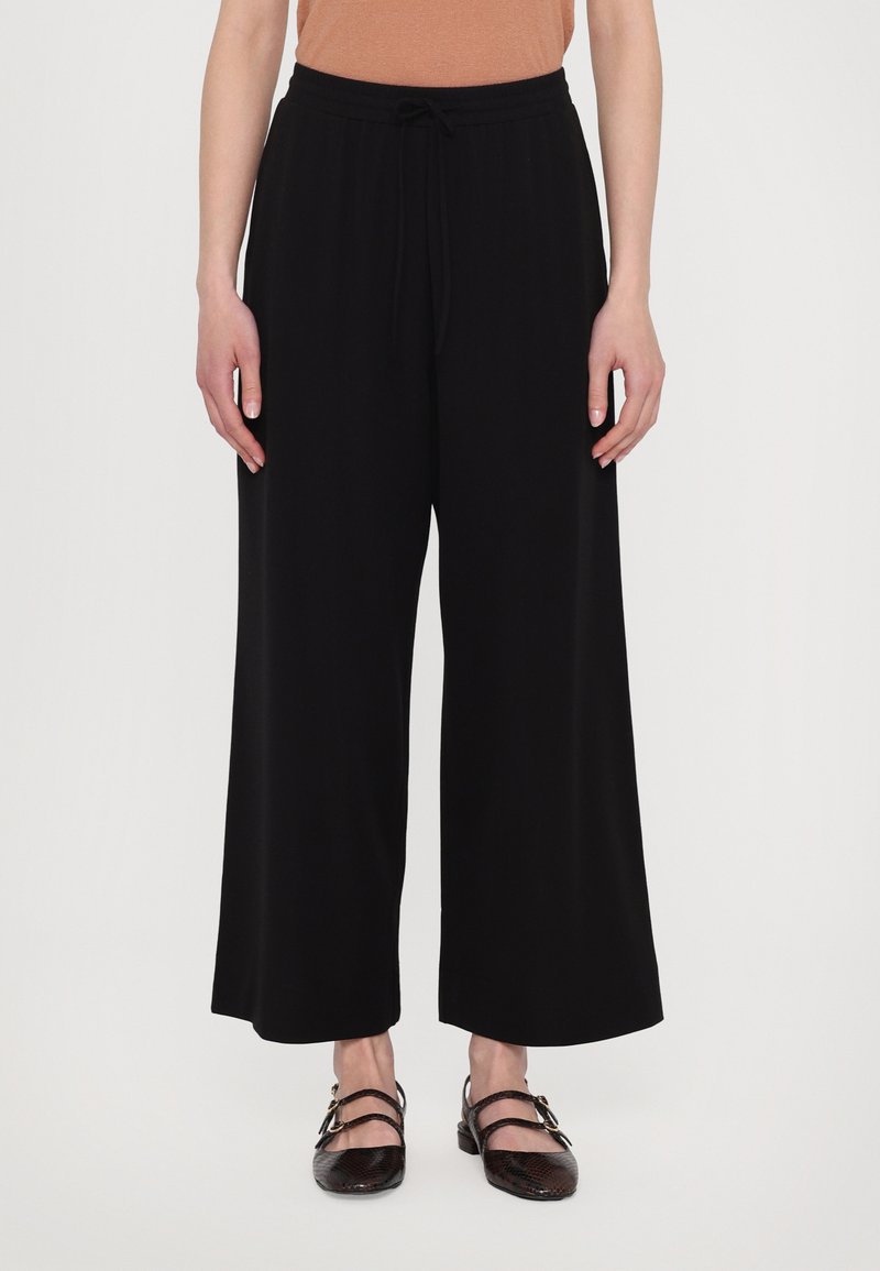 Black wide-leg pants made from a soft material with a drawstring waist. The model wears dark, textured shoes with buckle accents.