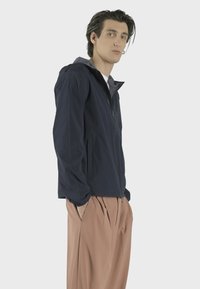 Námořnická lehká bunda s kapucí, vybavená zipovým uzavřením a bočními kapsami, v kombinaci s béžovými skládacími kalhotami. Hladká textura.