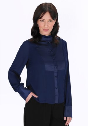 Sötét hajú nő, tengerészkék, hosszú ujjú, gombos blúzt és fekete nadrágot visel, egyik kezét a zsebében tartva áll.