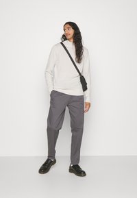 Maglia a maniche lunghe beige chiaro, pantaloni grigi e scarpe nere. Una piccola borsa a tracolla nera pende da una spalla. Sfondo semplice.