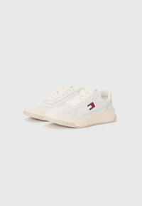 Vita sneakers med en slät läderoberdel, rund tå, platta snören och en beige gummisula. Har en liten logotyp i röd och blå färg.