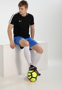 Zwarte Nike sportshirt met witte accenten, blauwe shorts, witte kniehoge sokken en zwarte voetbalschoenen, voet rustend op een felgekleurde voetbal.