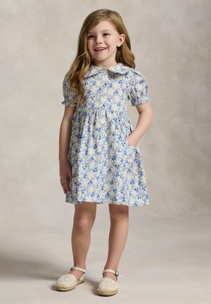 Νεαρό κορίτσι που χαμογελά, φοράει μπλε και άσπρο λουλουδάτο φόρεμα με φουσκωτά μανίκια και λευκά εσπαντρίγια, στέκεται με το ένα χέρι στην τσέπη.