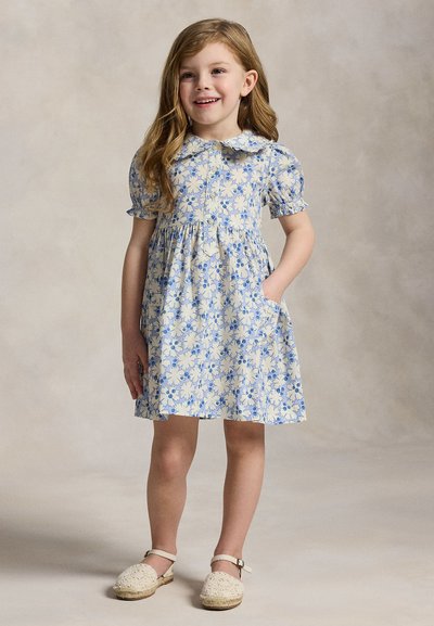 Νεαρό κορίτσι που χαμογελά, φοράει μπλε και άσπρο λουλουδάτο φόρεμα με φουσκωτά μανίκια και λευκά εσπαντρίγια, στέκεται με το ένα χέρι στην τσέπη.