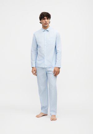 Homme debout pieds nus portant une chemise bleu clair à manches longues boutonnée et un pantalon bleu clair assorti sur un fond uni.