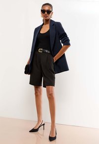 Kobieta w czarnym topie, granatowej marynarce z podwiniętymi rękawami, czarnych spodenkach z paskiem, czarnych butach na obcasie typu slingback, dużych kolczykach-kołach i okularach przeciwsłonecznych.