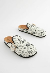 Baltas slip-on klēpju apavi ar melnu grafiku, korķa iekšzoles, regulējamu sprādzi un gumijas zole, ar zvaigžņu un sirds motīviem.